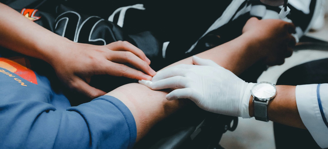 Mull wird von einer Hand mit Gummihandschuh in die Armbeuge einer anderen Person gedrückt, die andere Person, die man nur zum Tel sieht, drückt auch auf den Verband.