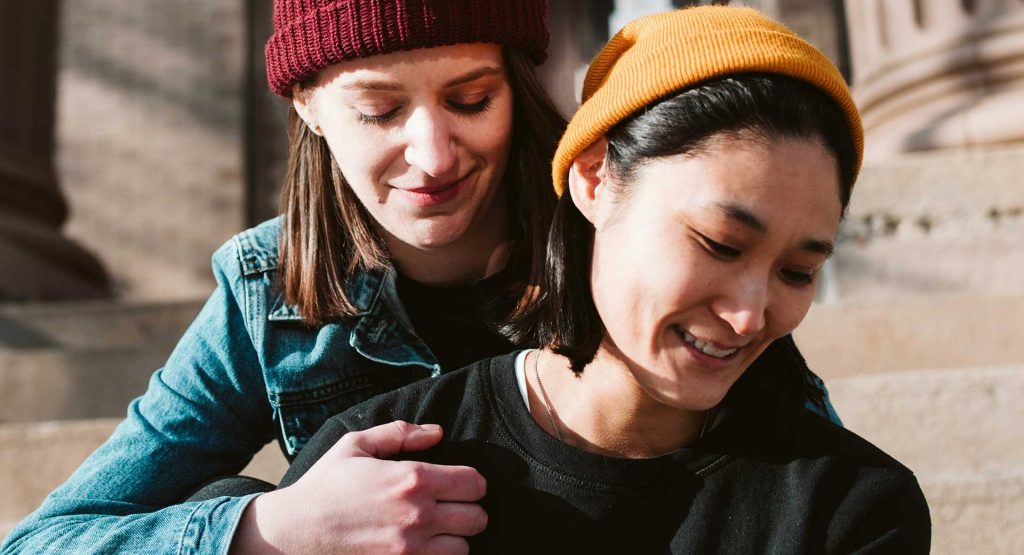 Profilansicht von zwei Frauen. Die hintere Frau fasst der vorderen Frau an die Schulter. Beide Frauen tragen Wollmützen.